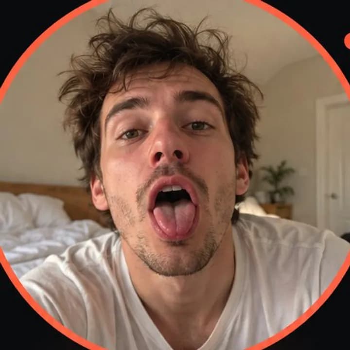 A sleepy man with messy hair pulling a face at his phone's front camera in a bedroom, mouth wide open and tongue out, framed in a coral circle.